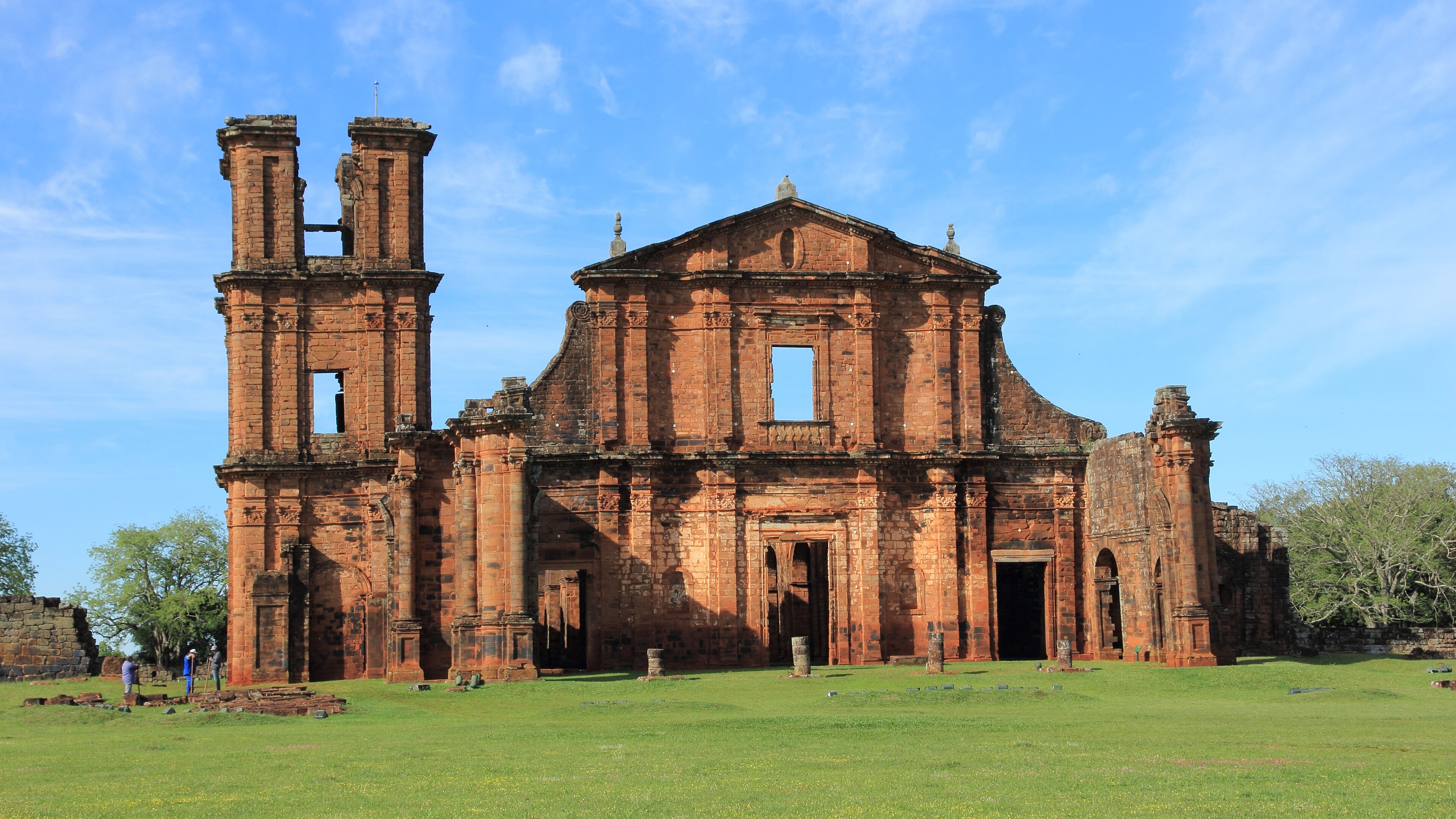 Fachada de arenito das ruínas da igreja de São Miguel Arcanjo, gramado e céu limpo.
